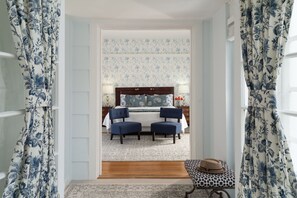 A bedroom with a double bed, two chairs, a small table with a hat, and floral curtains.