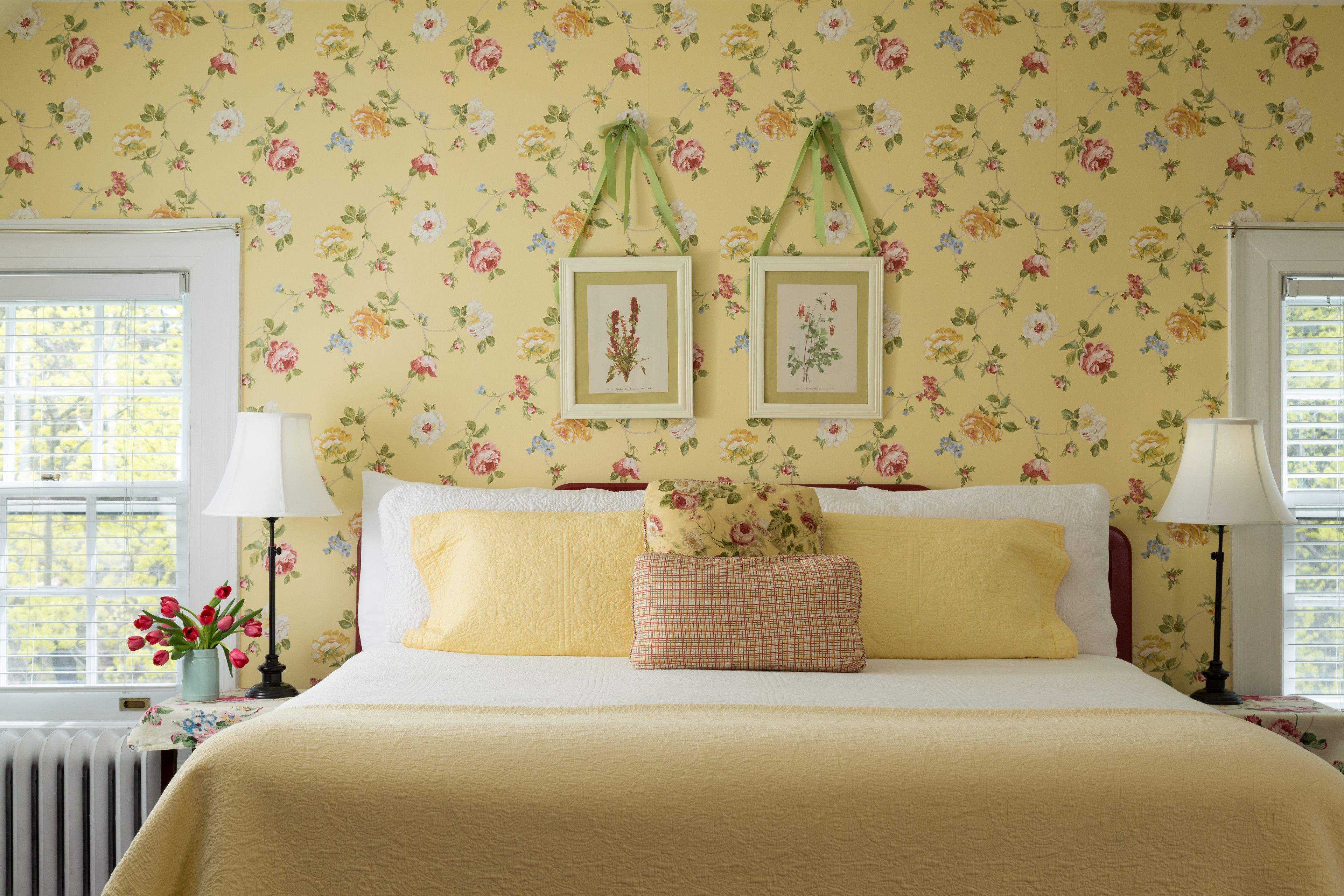 Ein Schlafzimmer mit floralem Wandpapier, einem Bett mit gelber Bettwäsche, zwei gerahmten botanischen Drucken an der Wand und einem Fenster mit Blick auf Bäume.