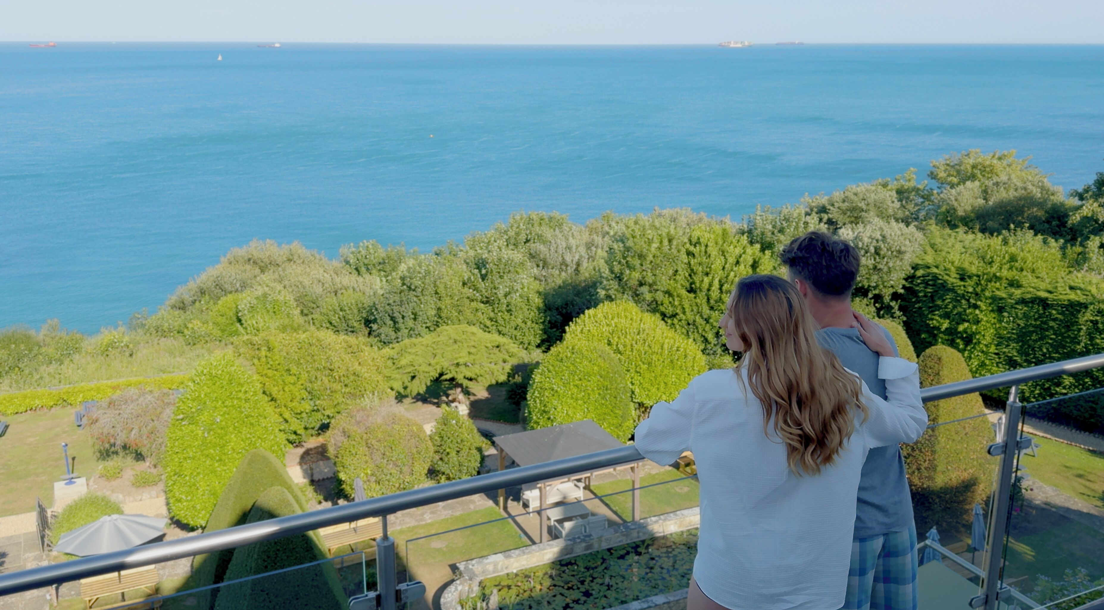 A couple overlooking a scenic view of the sea from a balcony with a garden and a small building in the background.