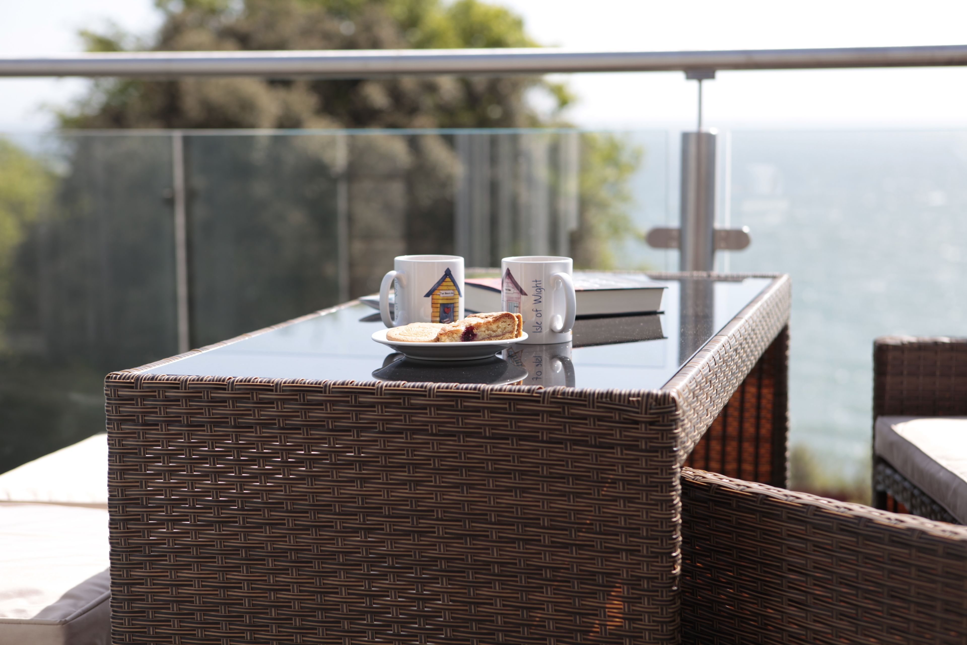 A wicker table with two mugs and a plate of food on a balcony overlooking a body of water.