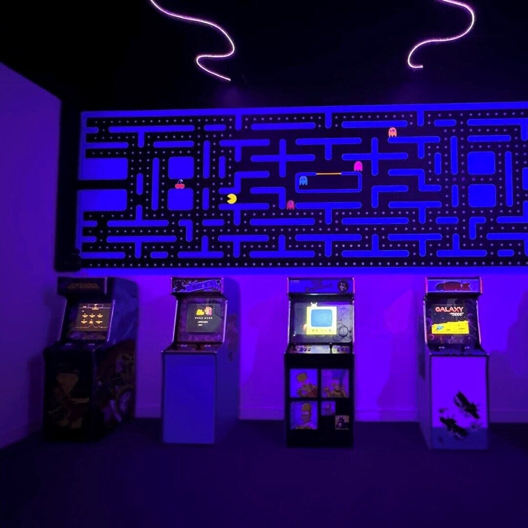 Une pièce dont les murs sont décorés de motifs complexes évoquant un labyrinthe, avec des jeux d’arcade et des néons.