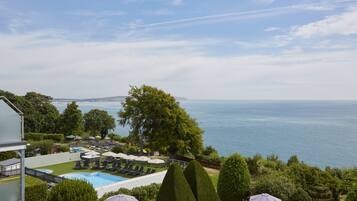 A pool area with lounge chairs and umbrellas, surrounded by greenery and overlooking a body of water.