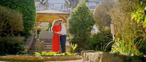 A couple embracing in a garden with stone steps, a wooden pergola, and a variety of plants.