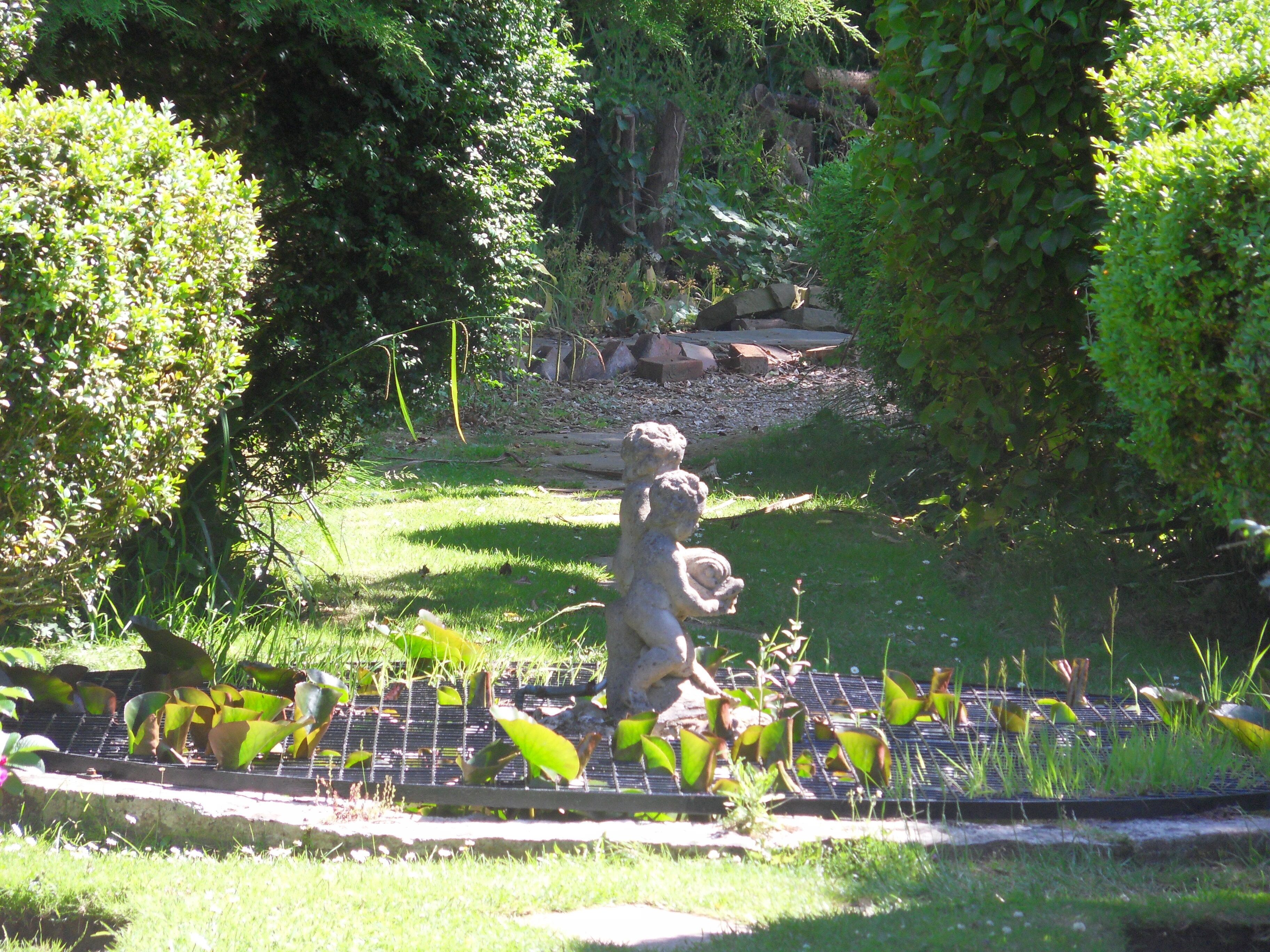 A garden with a stone statue, a pond with water lilies, and a grassy area.