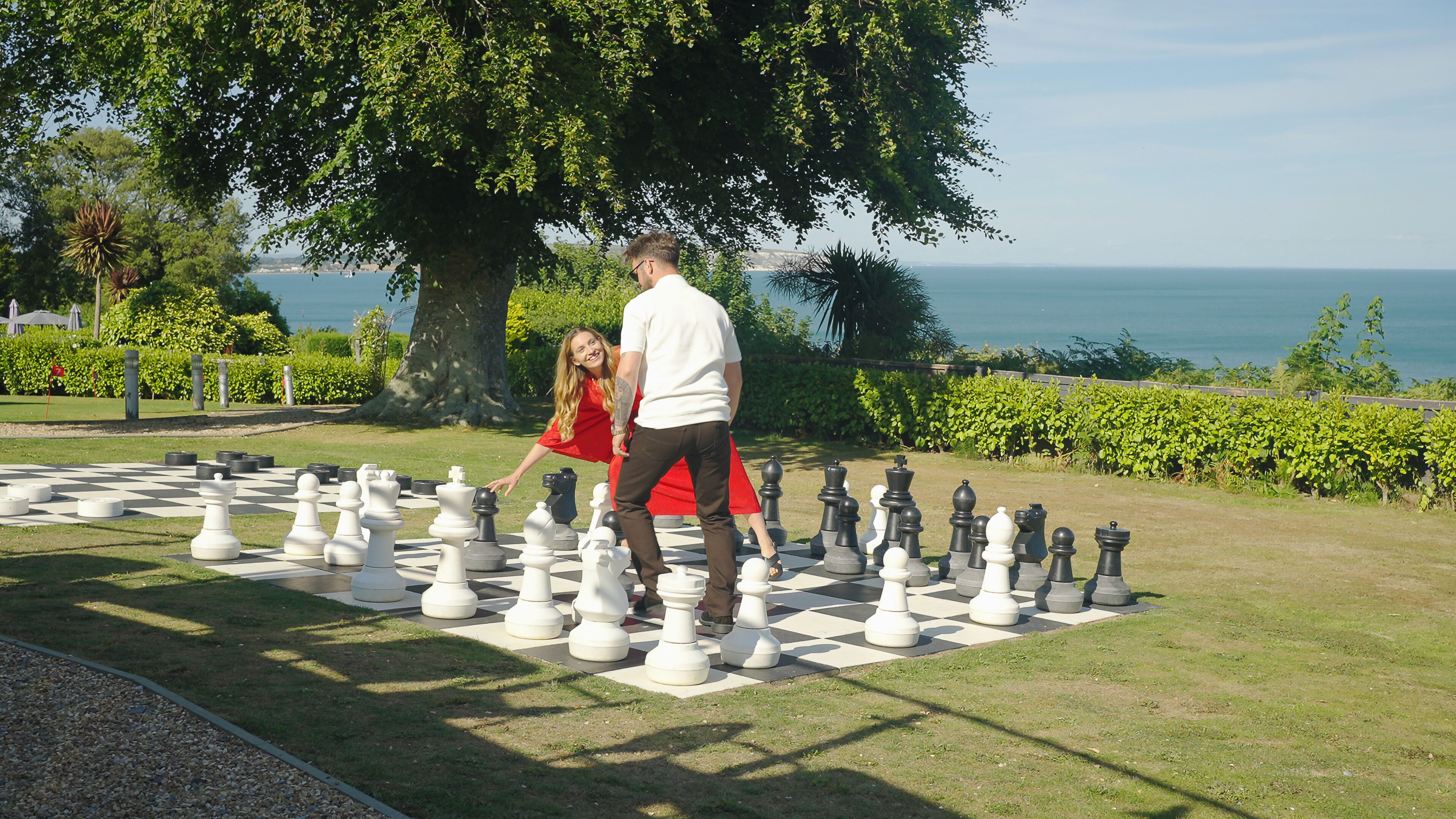 A large outdoor chessboard set up on a grassy area with a view of the sea in the background.