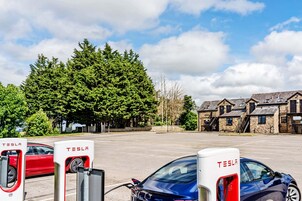 Una estación de carga Tesla con dos coches, uno rojo y otro azul, en un aparcamiento con un edificio de piedra y árboles al fondo.