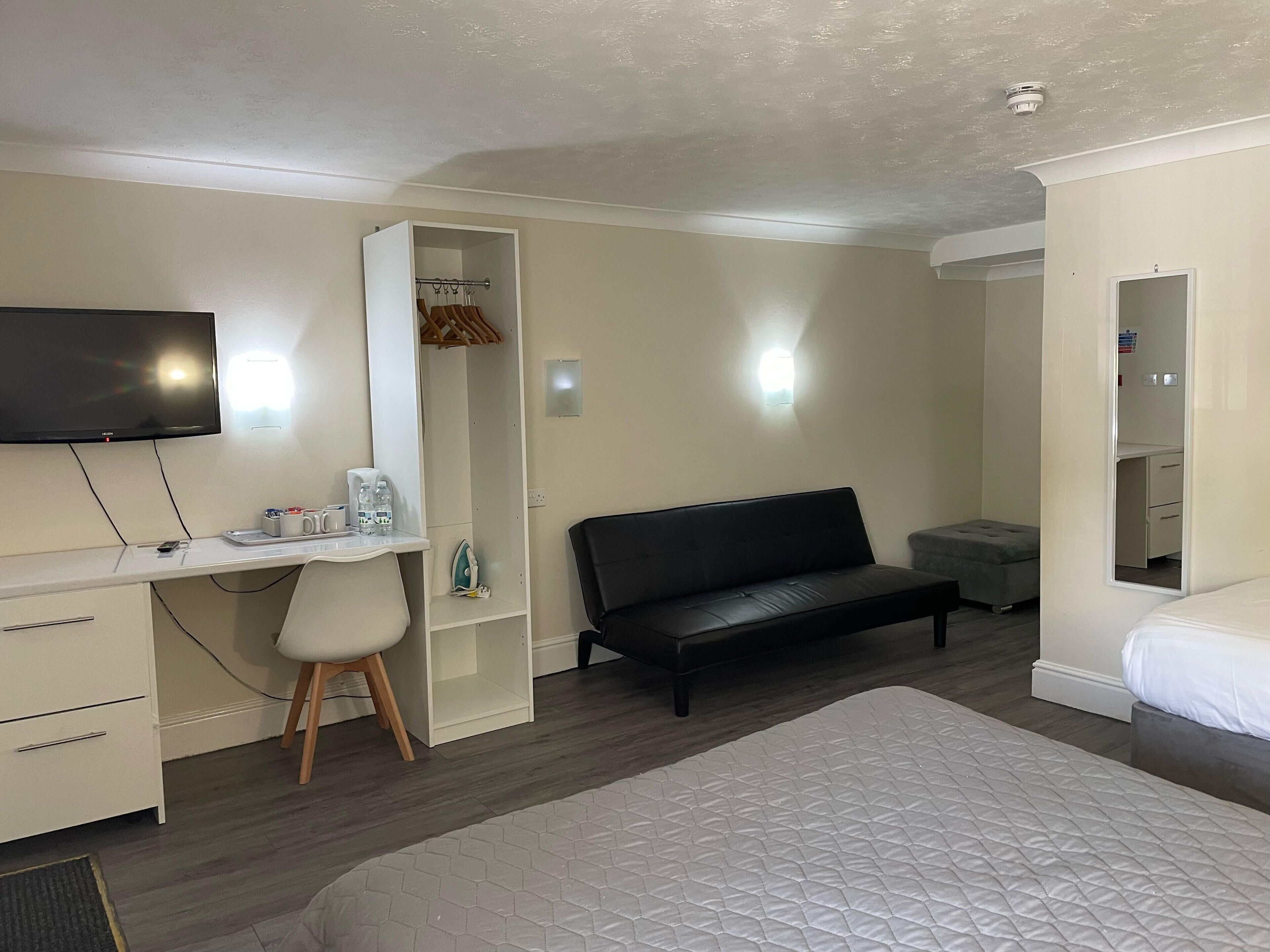 A hotel room with a bed, a desk with a chair, a black sofa, a wardrobe, a television, and a small table with a coffee maker and water bottles.