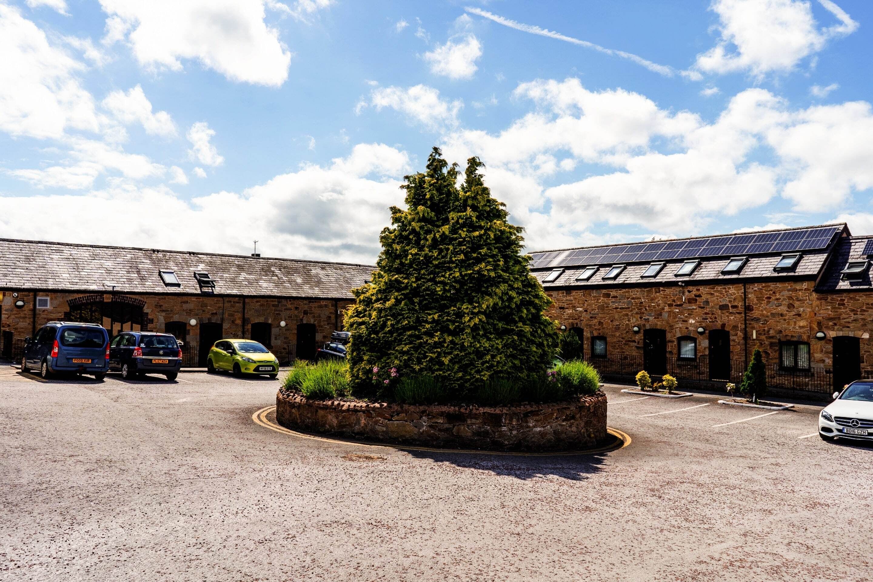 A building with a gravel parking area, a large evergreen tree, and solar panels on the roof.