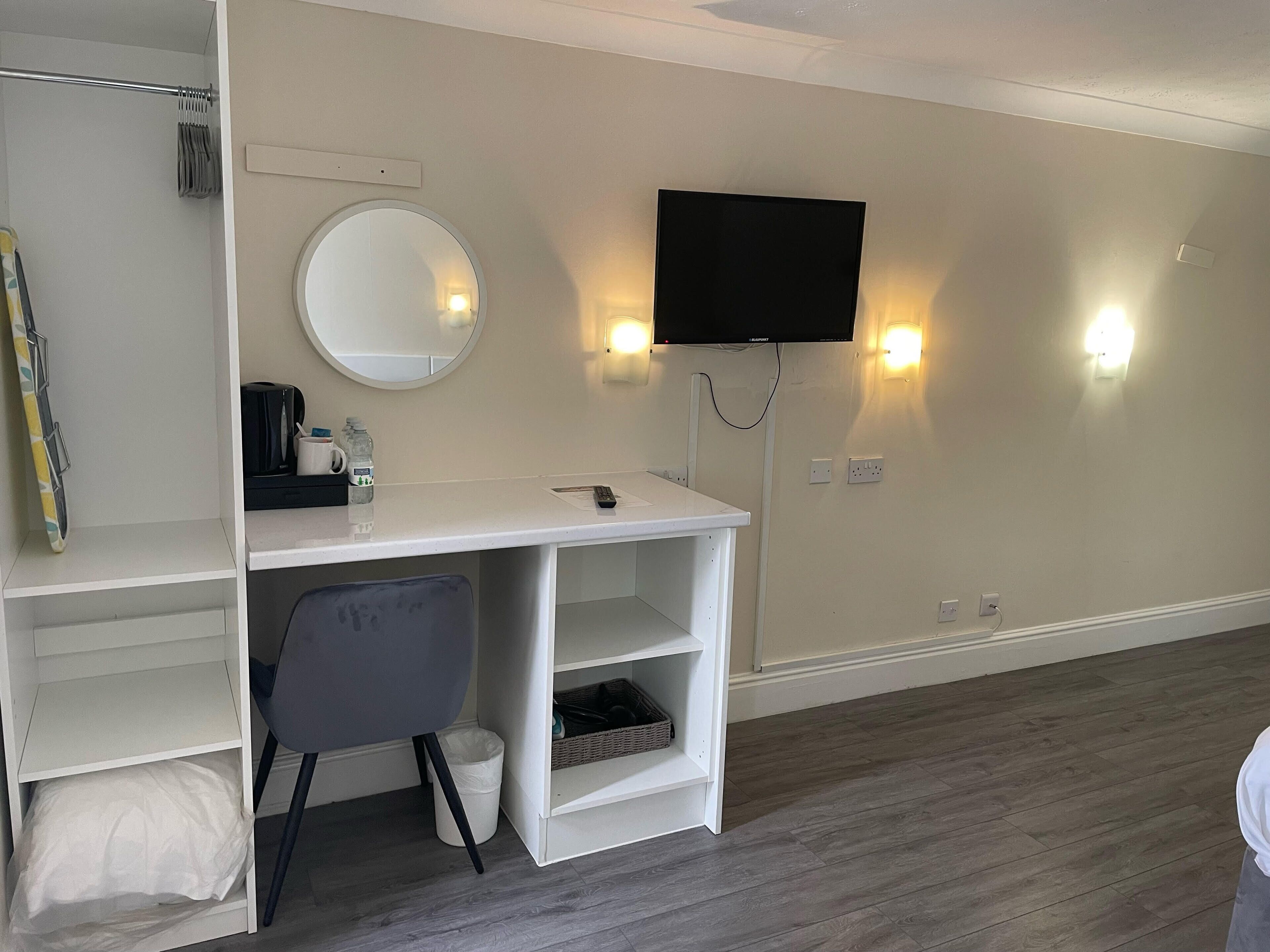 A hotel room with a white desk, a chair, a round mirror, a flat-screen TV, and a shelf with a basket.