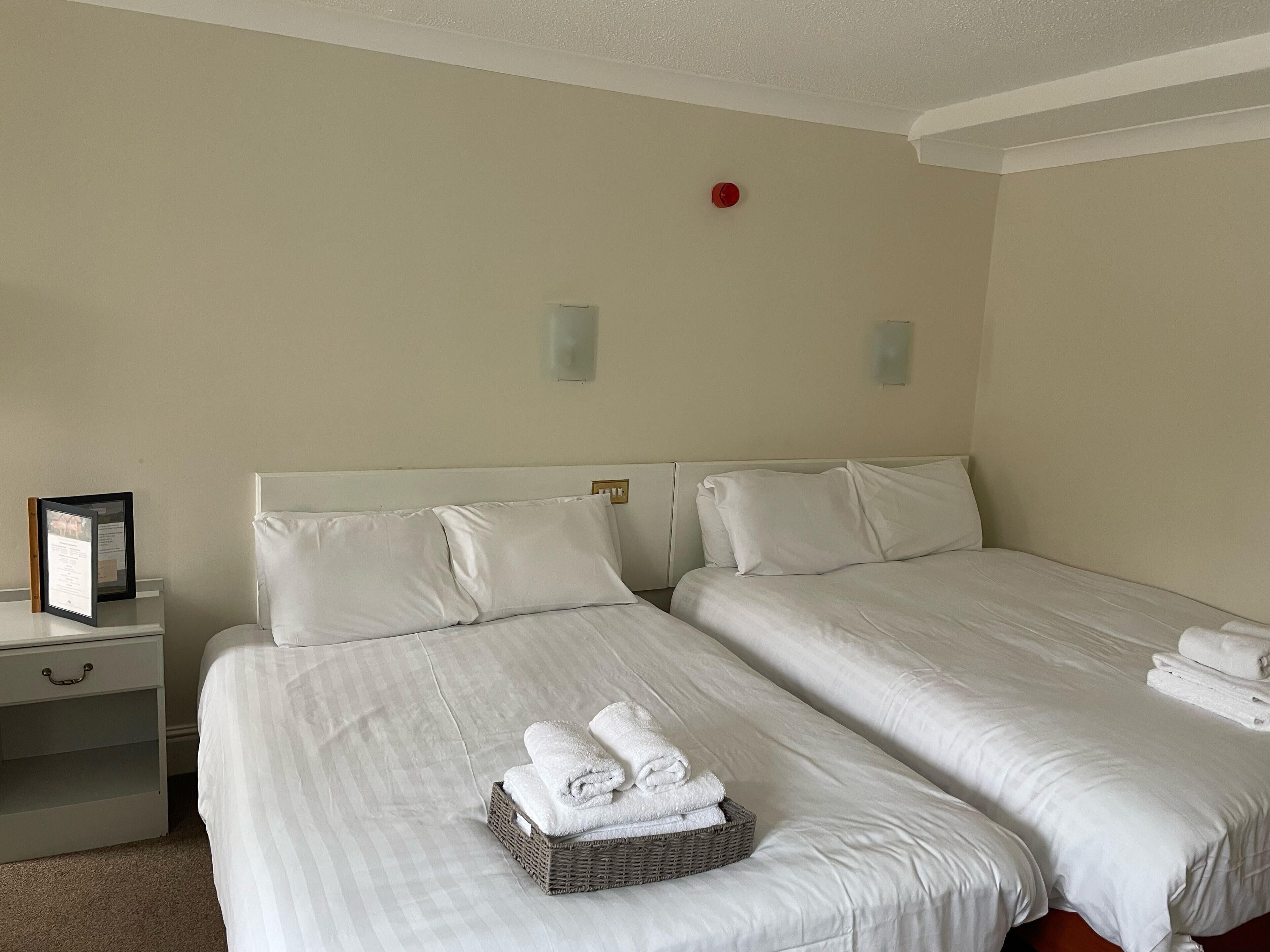 A hotel room with two single beds, a nightstand with a framed picture, and a basket of folded towels.