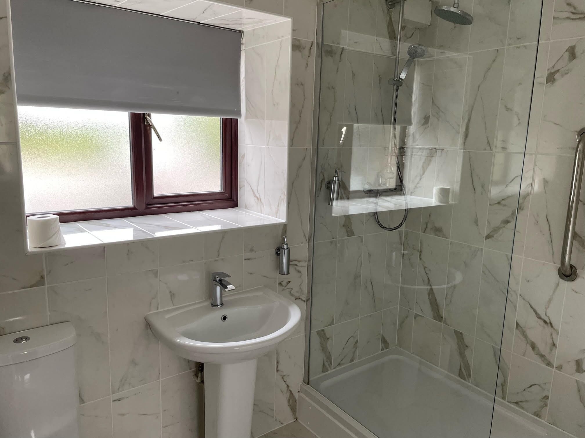 A bathroom with a marble-tiled shower, a sink, and a window with a blind.