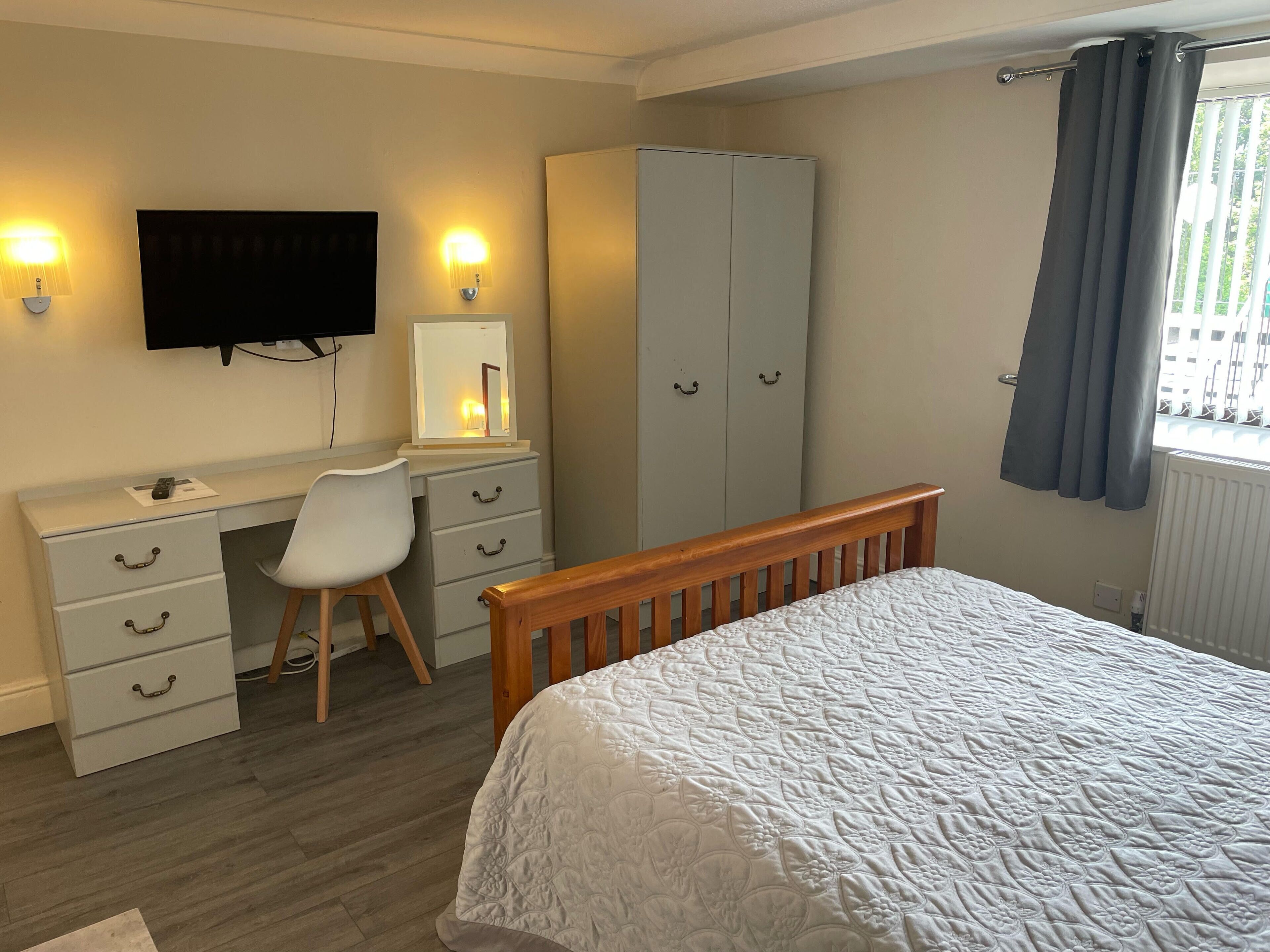 Ein Schlafzimmer mit einem Bett, einem Schreibtisch mit Stuhl, einem Kleiderschrank, einem Fernseher und einem Fenster mit Vorhängen.
