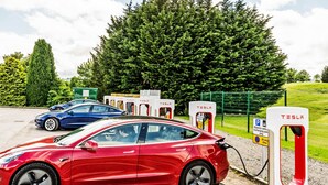 A red Tesla car parked at a charging station with a green fence and trees in the background. - Mountain Park Hotel (Flint)