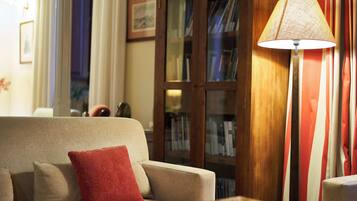 Een gezellige woonkamer met een beige bank, een rode kussen, een vloerlamp en een boekenplank vol boeken.