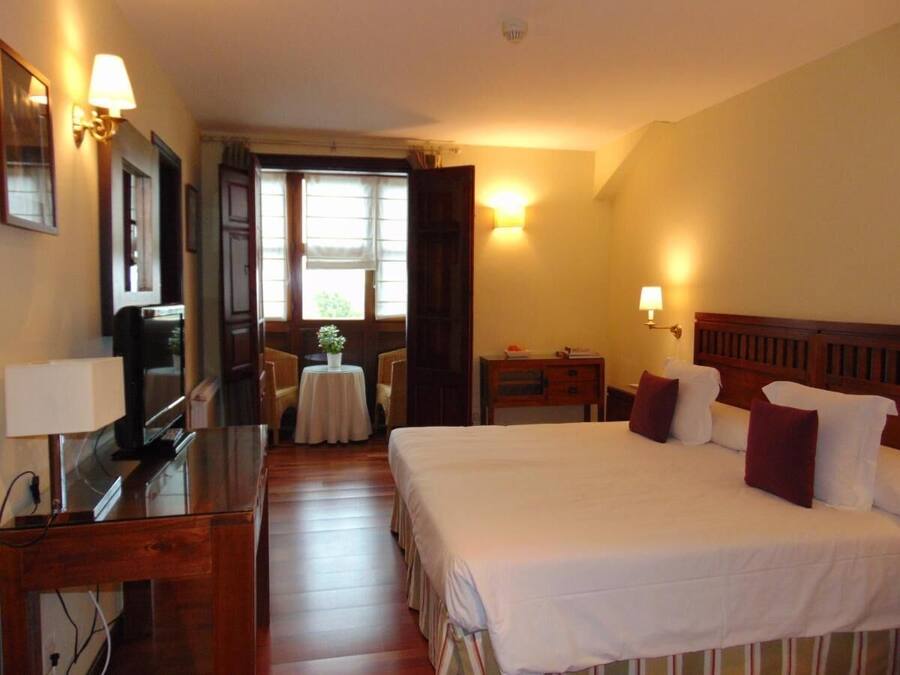 A hotel room with a bed, bedside table, lamp, desk, and a small table with a plant.
