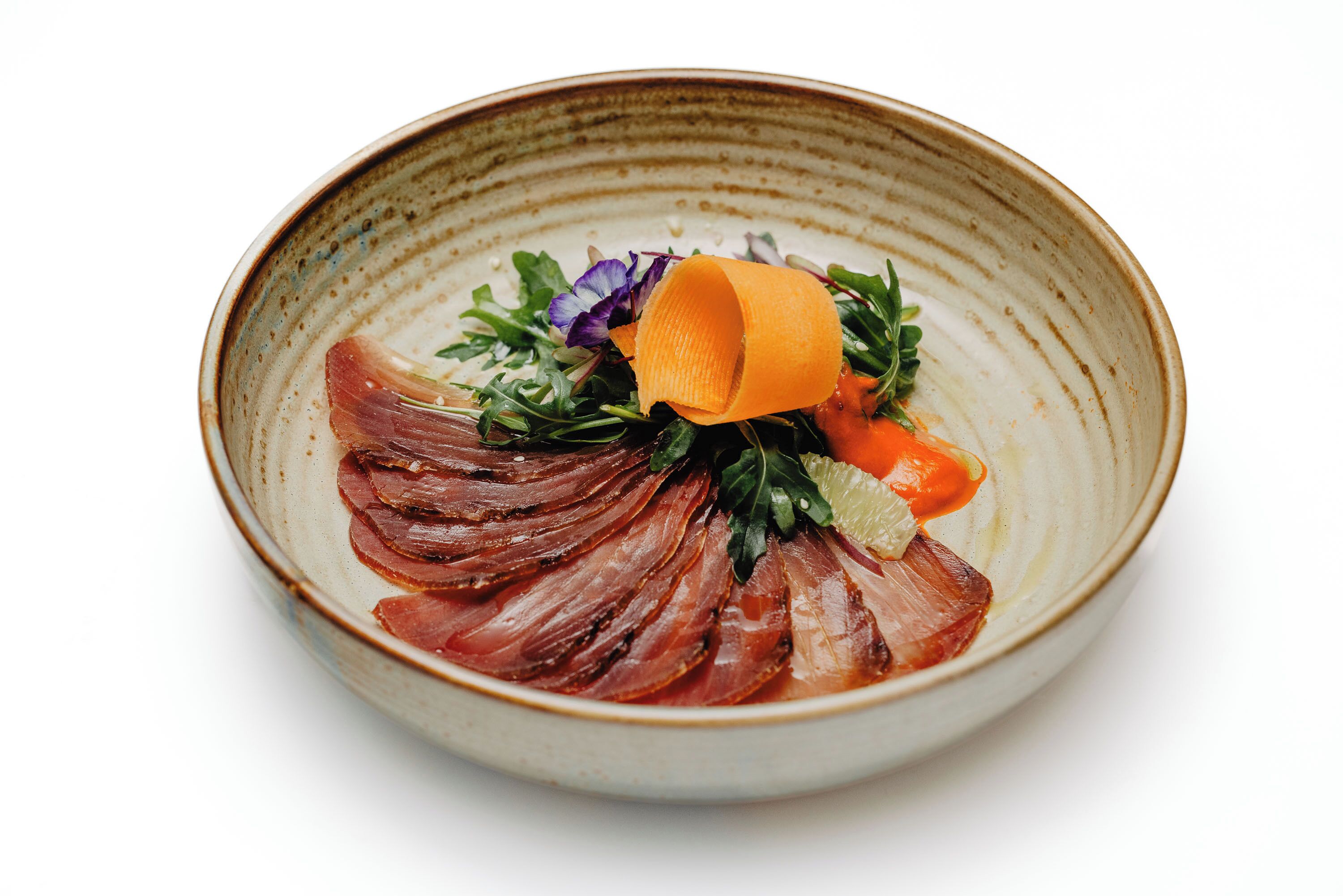 A ceramic bowl with a layered food presentation, including thinly sliced meat, greens, and a small orange element.