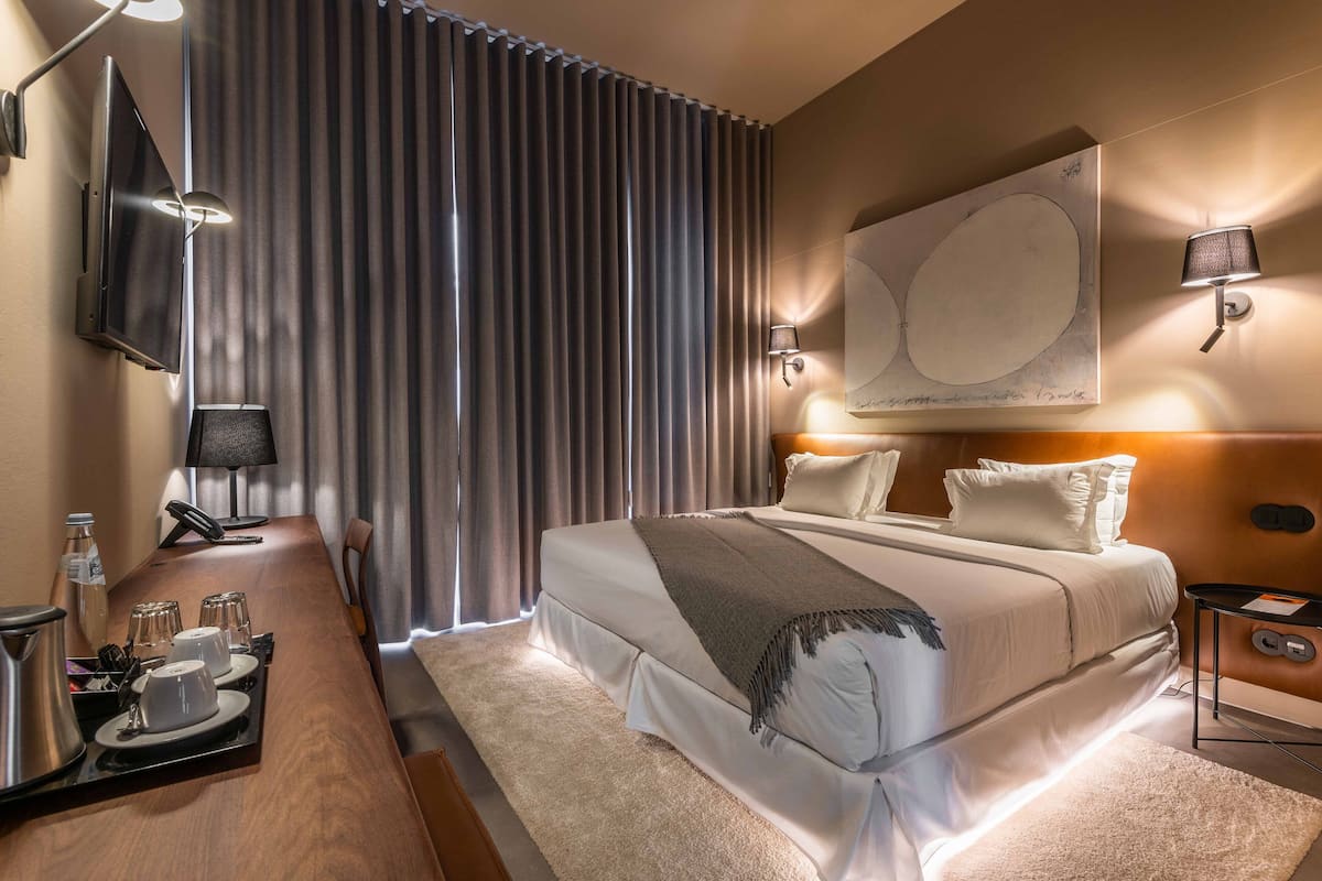 Une chambre d’hôtel avec un grand lit, une table de chevet avec une lampe, un téléviseur à écran plat fixé au mur et une table à manger dressée avec des verres et une cafetière.