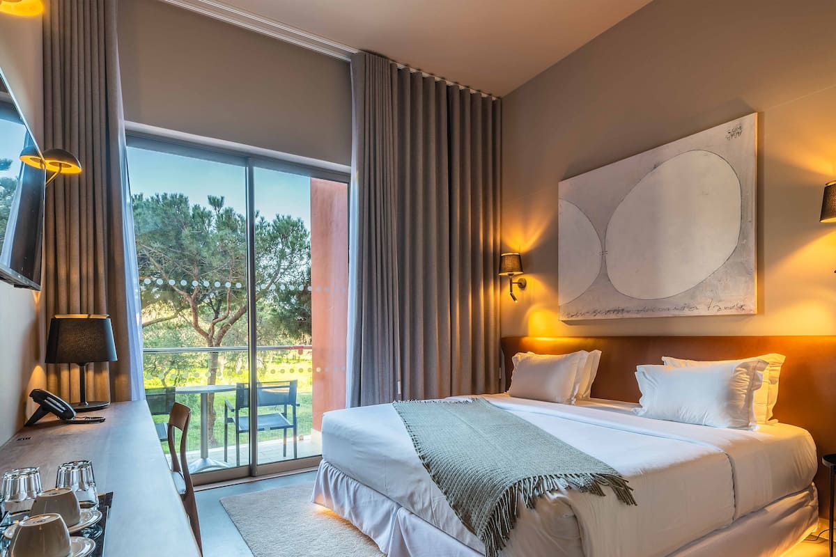 Une chambre d’hôtel avec un grand lit, un bureau avec une lampe et une vue sur les arbres par la fenêtre.
