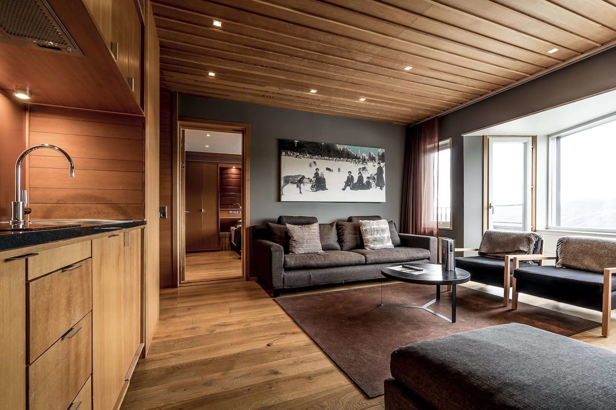 Ein modernes Wohnzimmer mit Holzboden, einem grauen Sofa, einem Couchtisch und Blick auf eine verschneite Landschaft durch große Fenster.