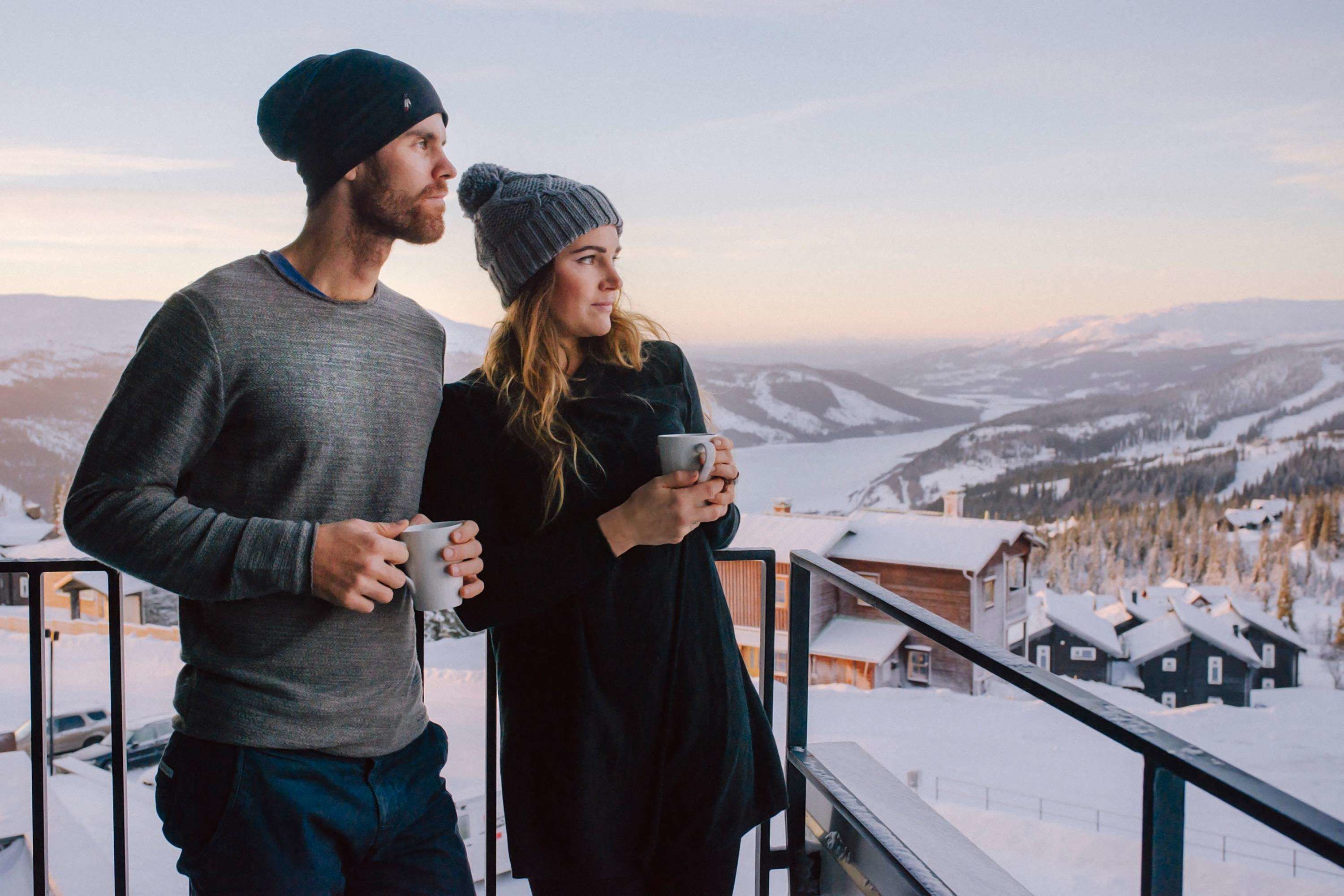 Un couple se tient sur un balcon, d’où l’on peut admirer un paysage enneigé avec des maisons en arrière-plan.