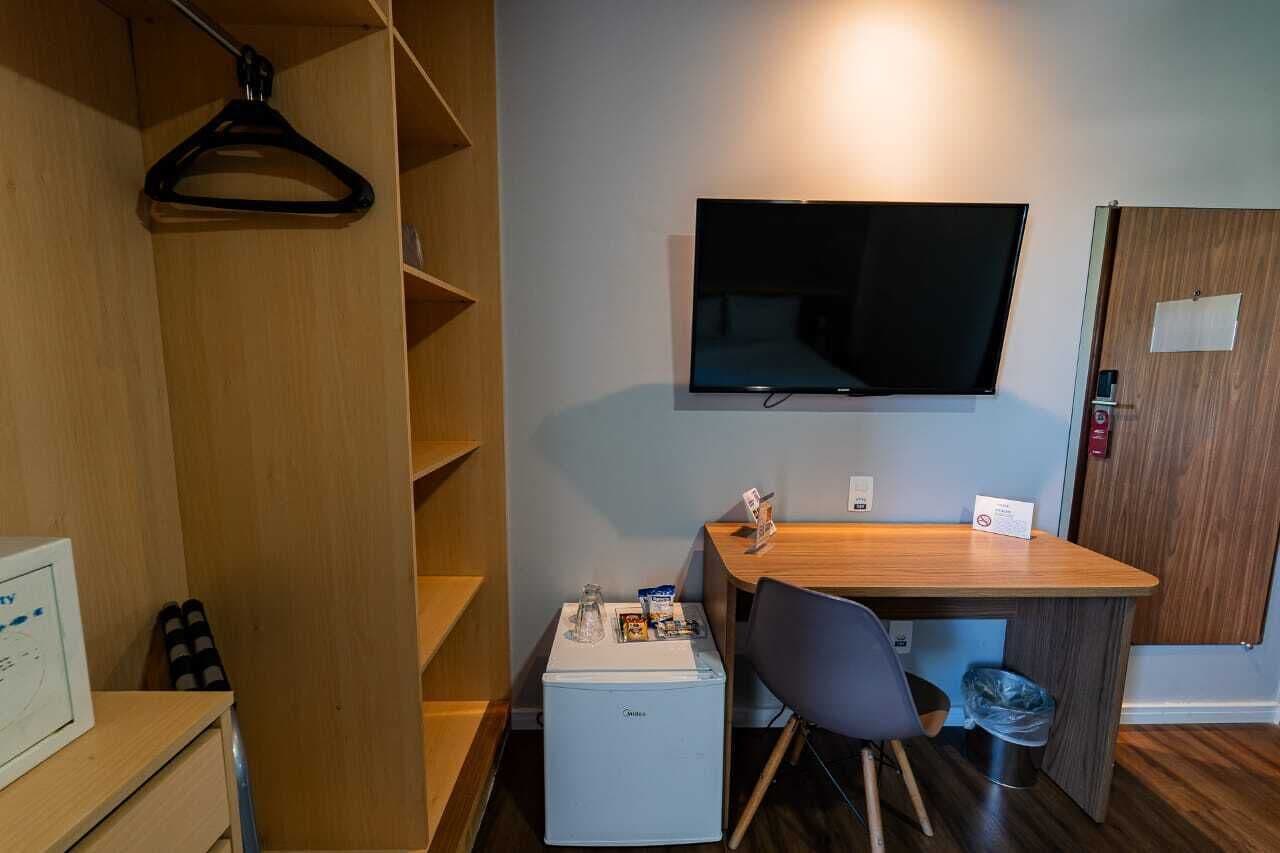 A compact room with a flat-screen TV mounted on the wall, a small wooden desk with a chair, a mini-fridge, and a coat rack.
