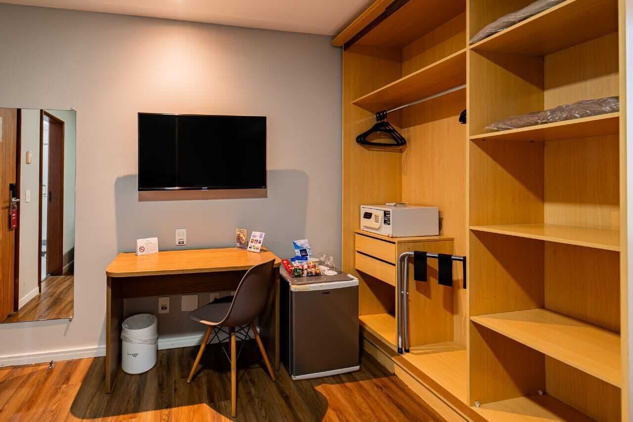 Habitación compacta con escritorio, silla, un refrigerador pequeño, microondas y televisión montada en la pared.