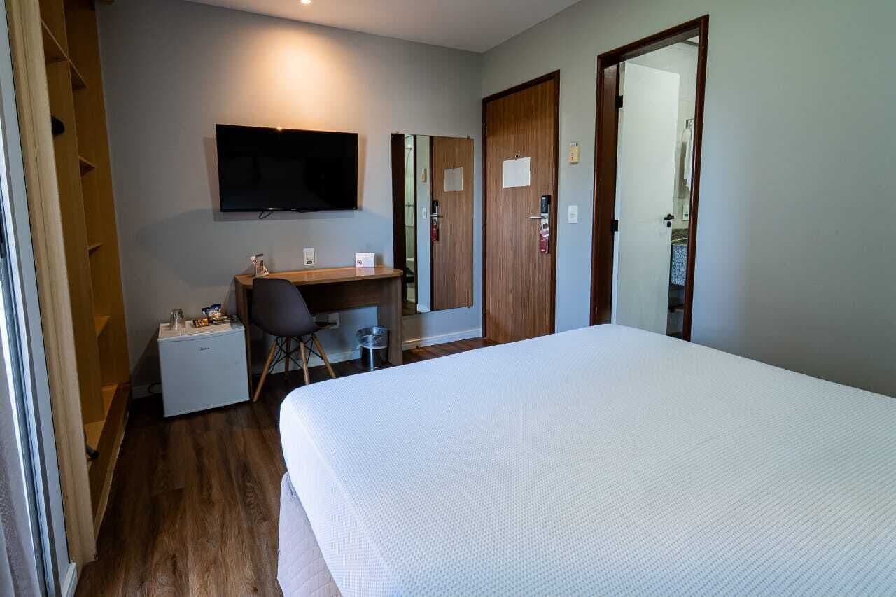Quarto de hotel com cama, escrivaninha, televisão e guarda-roupa.