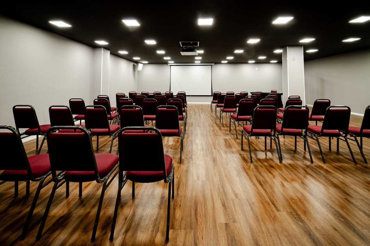 Konferenssihuone, jossa on riveissä punaisia tuoleja, projektorikangas ja puulattia.