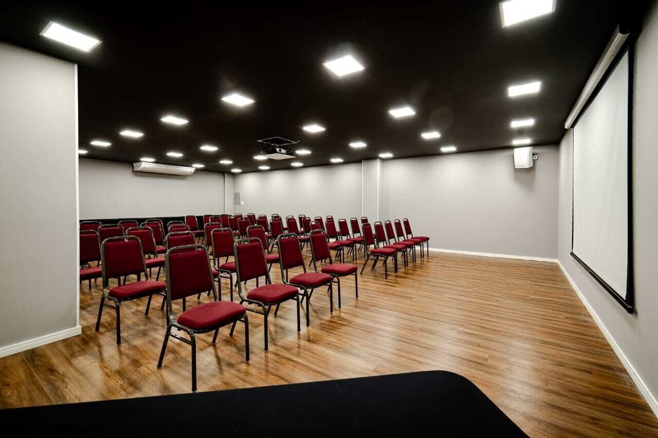 Un salón de conferencias con hileras de sillas rojas, una pantalla de proyector e iluminación en el techo.