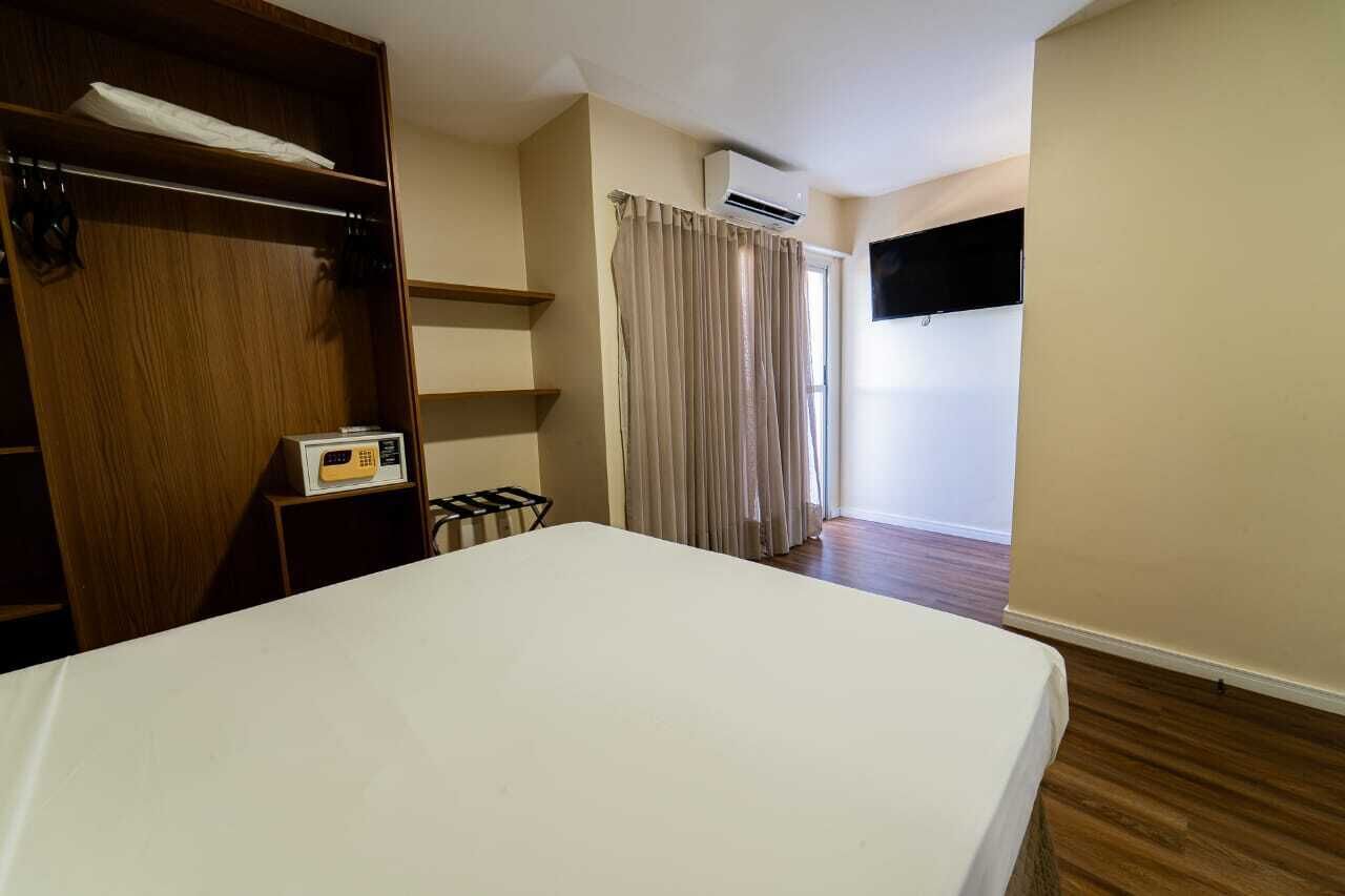 A bedroom with a bed, a wardrobe, a small table with a radio, a window with curtains, and an air conditioning unit.