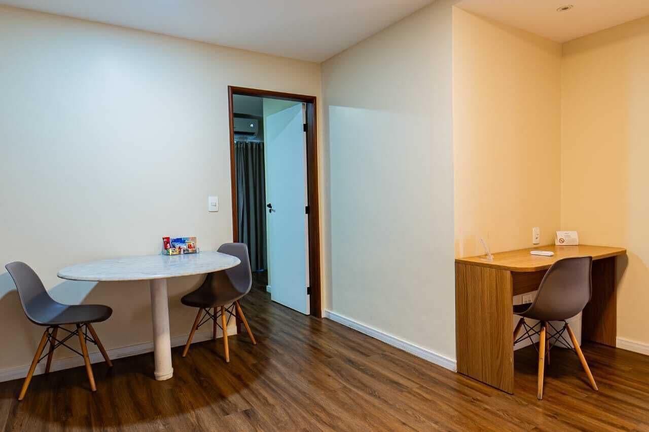 Quarto com uma mesa redonda e duas cadeiras, uma pequena escrivaninha com uma cadeira e uma porta que dá para outro cômodo.