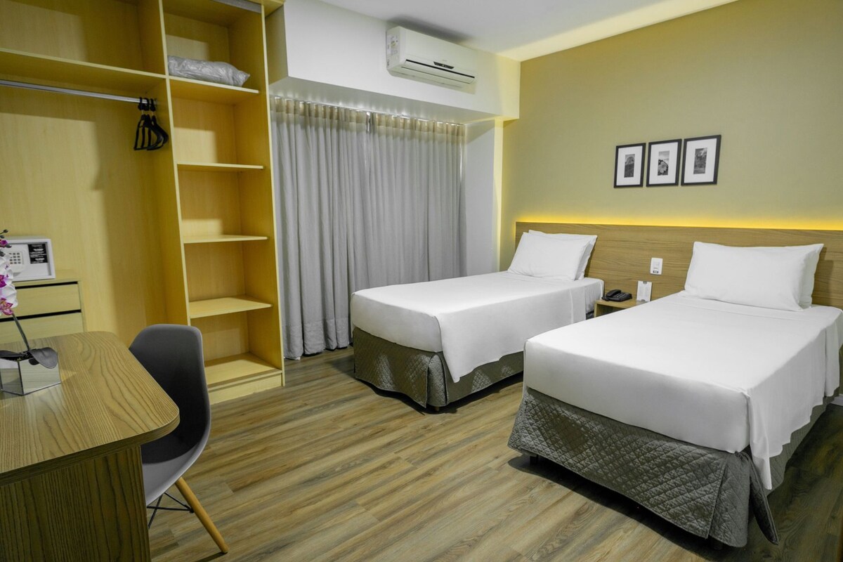 A hotel room with two beds, a desk, a chair, a wardrobe, and an air conditioning unit.
