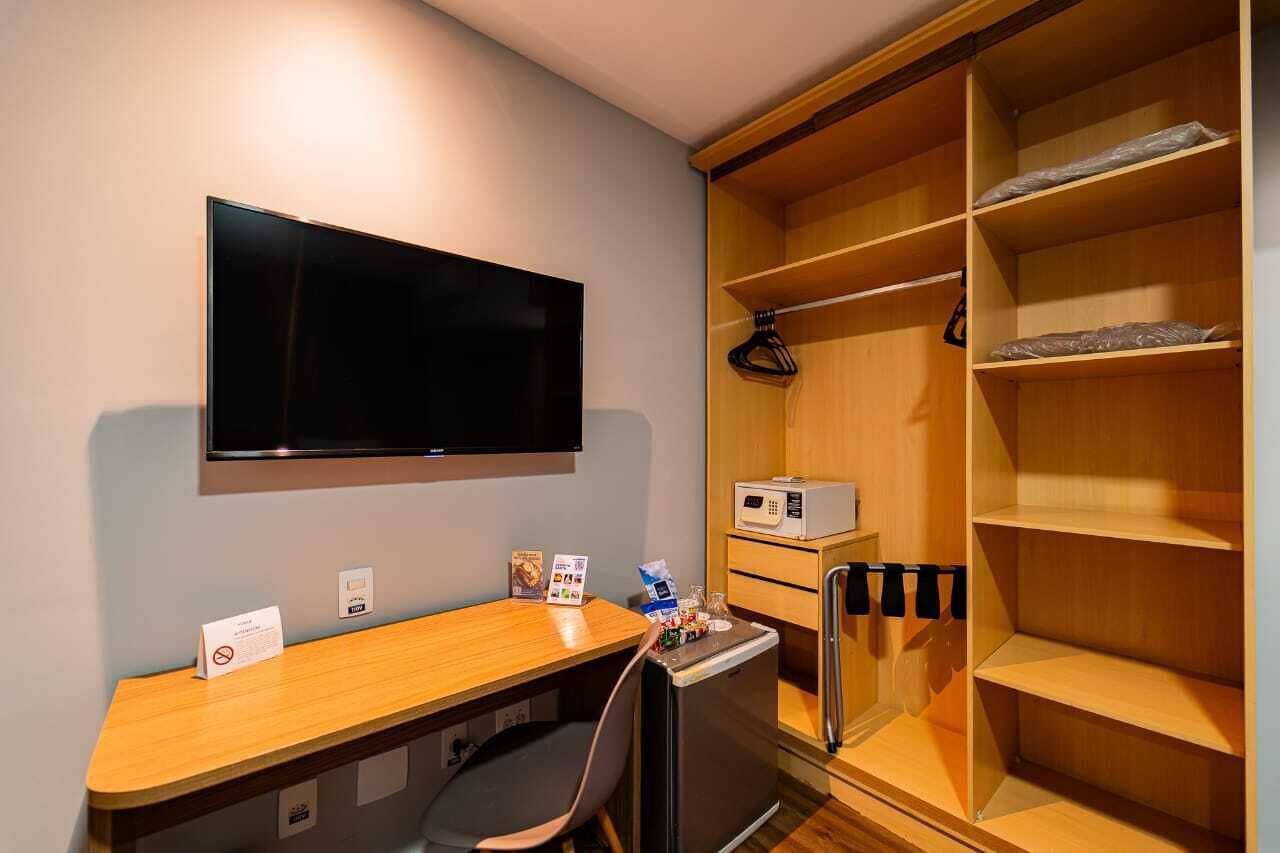 A room with a desk, a television, a mini-fridge, a microwave, and a wooden wardrobe with shelves.