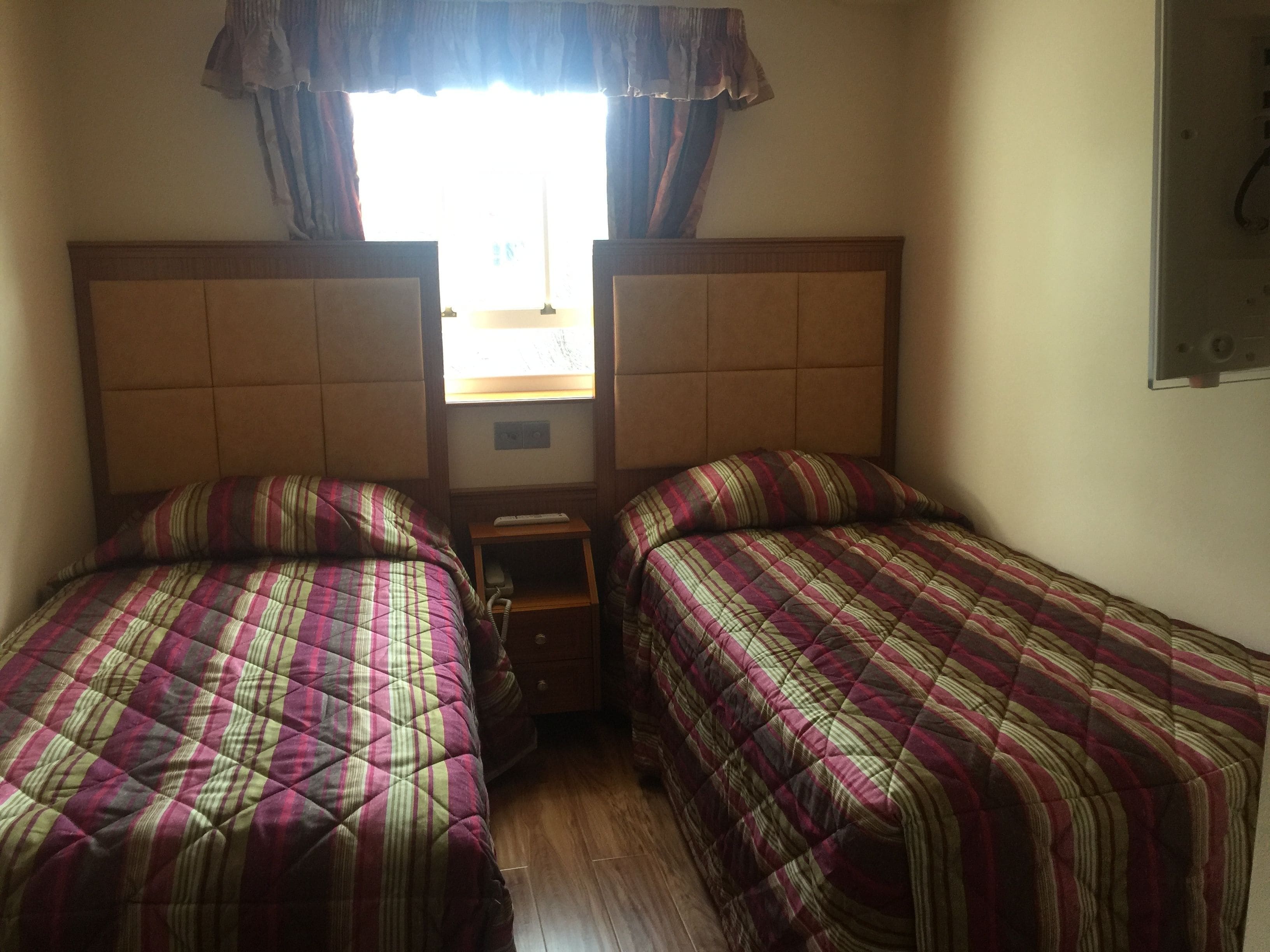 A room with two beds, each with a striped bedspread, a small wooden nightstand, and a window with curtains.