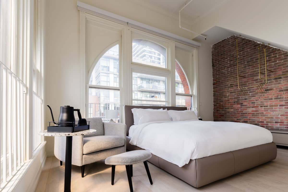 Une chambre moderne avec un grand lit, une petite table avec une bouilloire, une chaise et une vue sur les bâtiments à travers des fenêtres en arc de cercle.