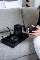 Iemand houdt een mok vast, zittend op een grijze bank met een zwarte dienblad waarop een koffiepot en een kopje staan.