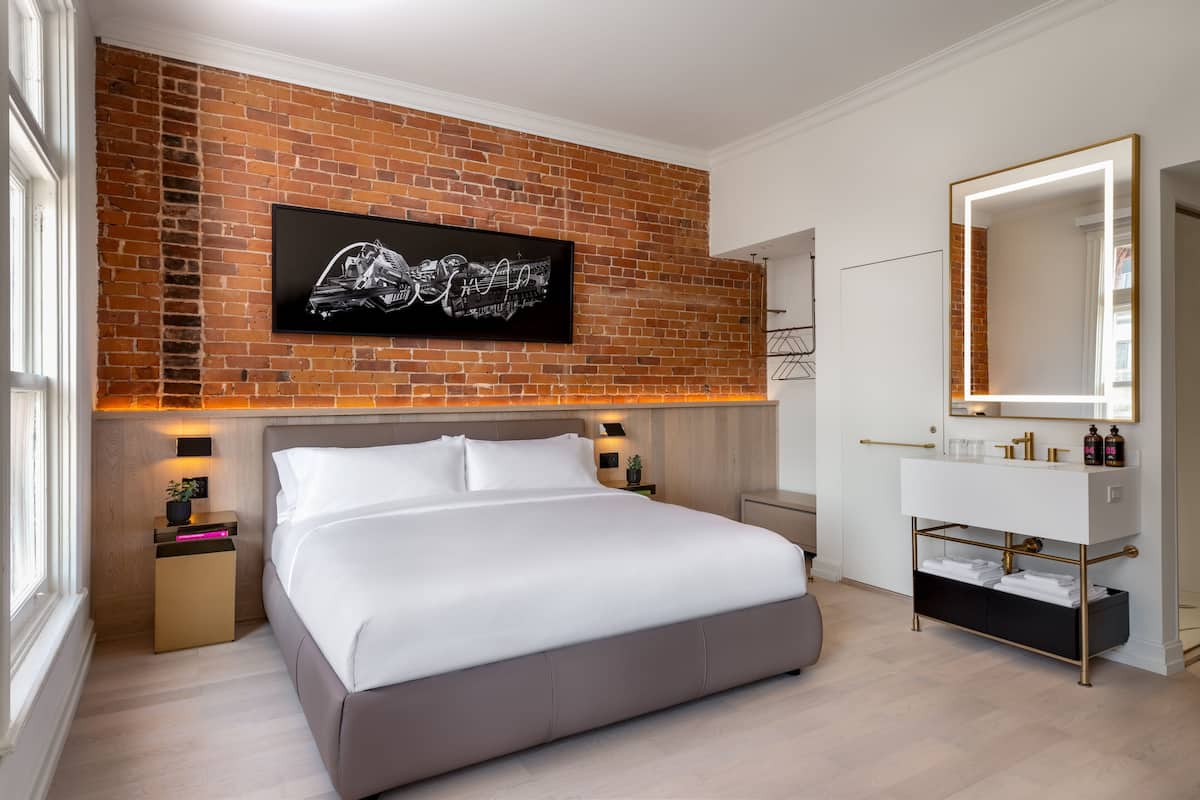 Une chambre à coucher moderne avec un grand lit, un mur d’accent en briques, un téléviseur fixé au mur, une table de chevet avec une lampe et une coiffeuse avec un miroir.