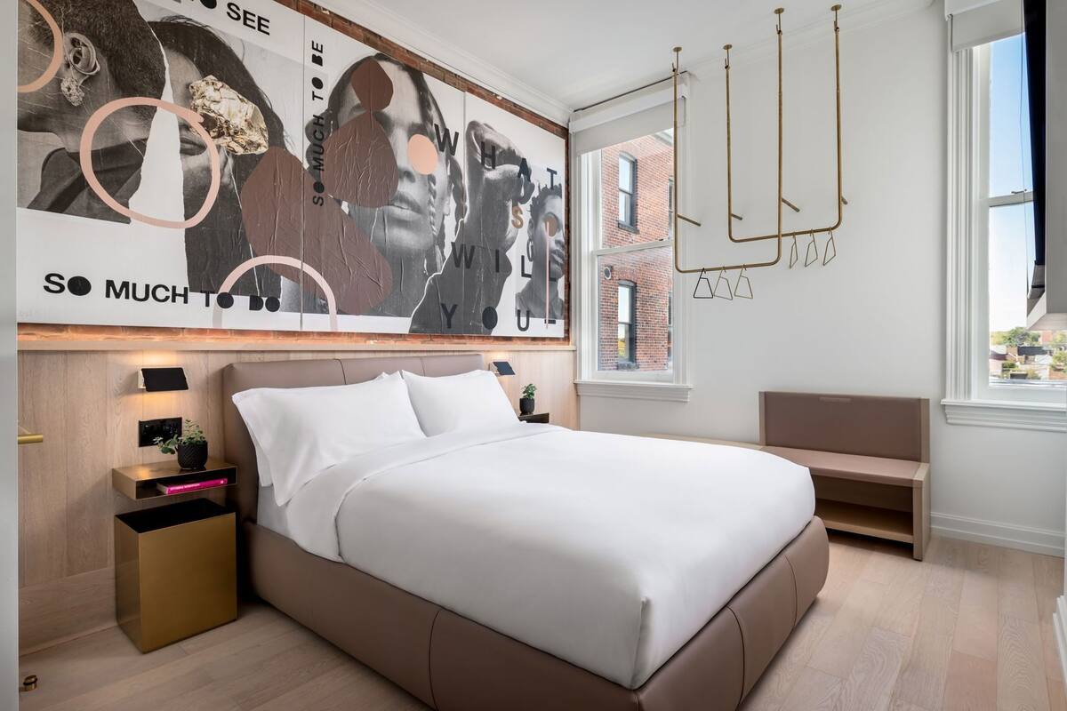 Une chambre à coucher moderne avec un grand lit, des tables de chevet, un banc et une fresque représentant un paysage urbain sur le mur.