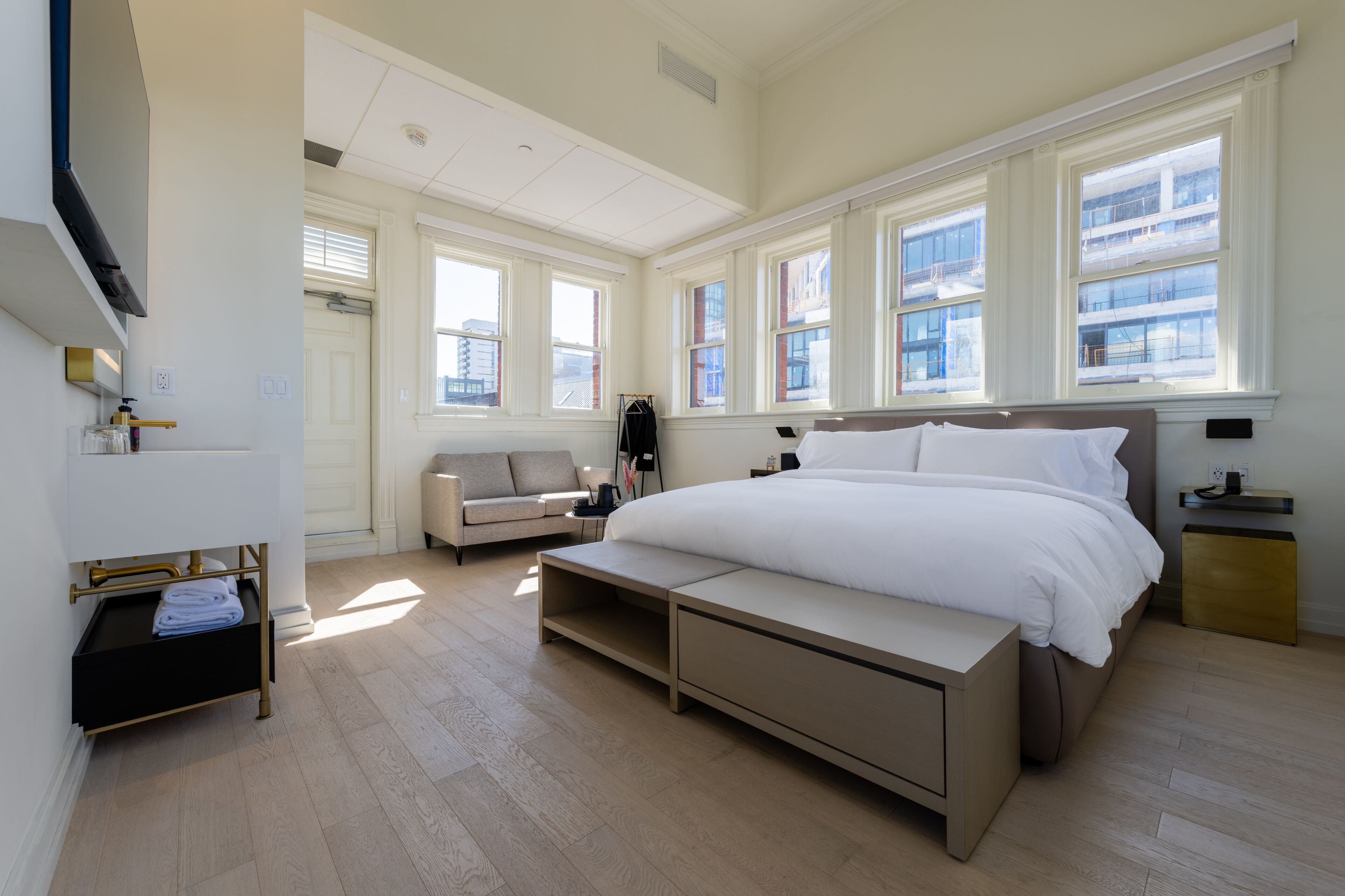 Un dormitorio moderno con una cama grande, un sofá y un escritorio. Hay varias ventanas que dejan entrar luz natural y ofrecen vistas de la ciudad.