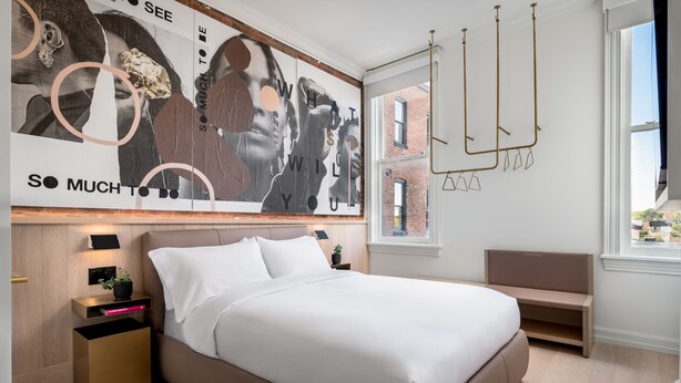 A modern bedroom with a large bed, a bedside table with a lamp, a wall-mounted mirror, and a window with a view of buildings.