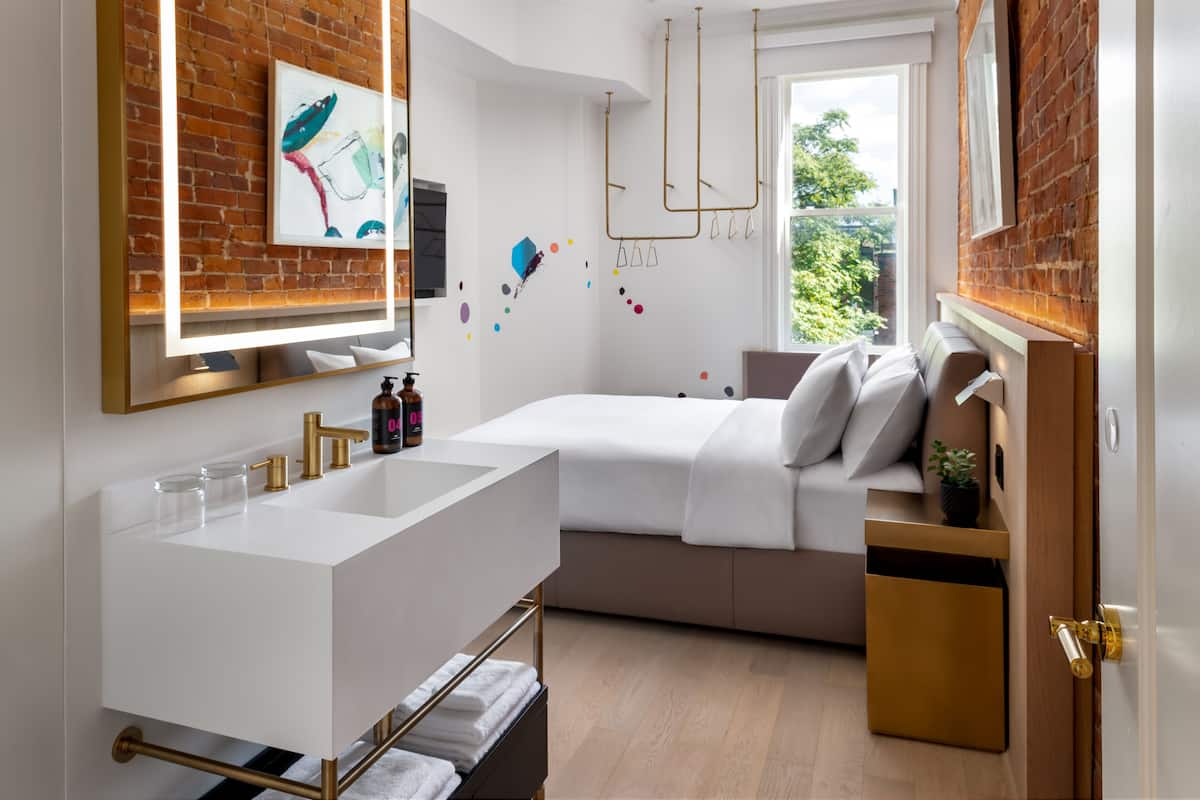 Une salle de bain moderne avec un grand lavabo blanc, un miroir et un espace douche. On y trouve un lit avec une tête de lit blanche et une table de chevet agrémentée d’une plante.