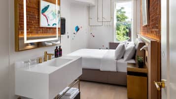 A modern bathroom with a large white sink, a mirror, and a shower area. There is a bed with a white headboard and a bedside table with a plant.