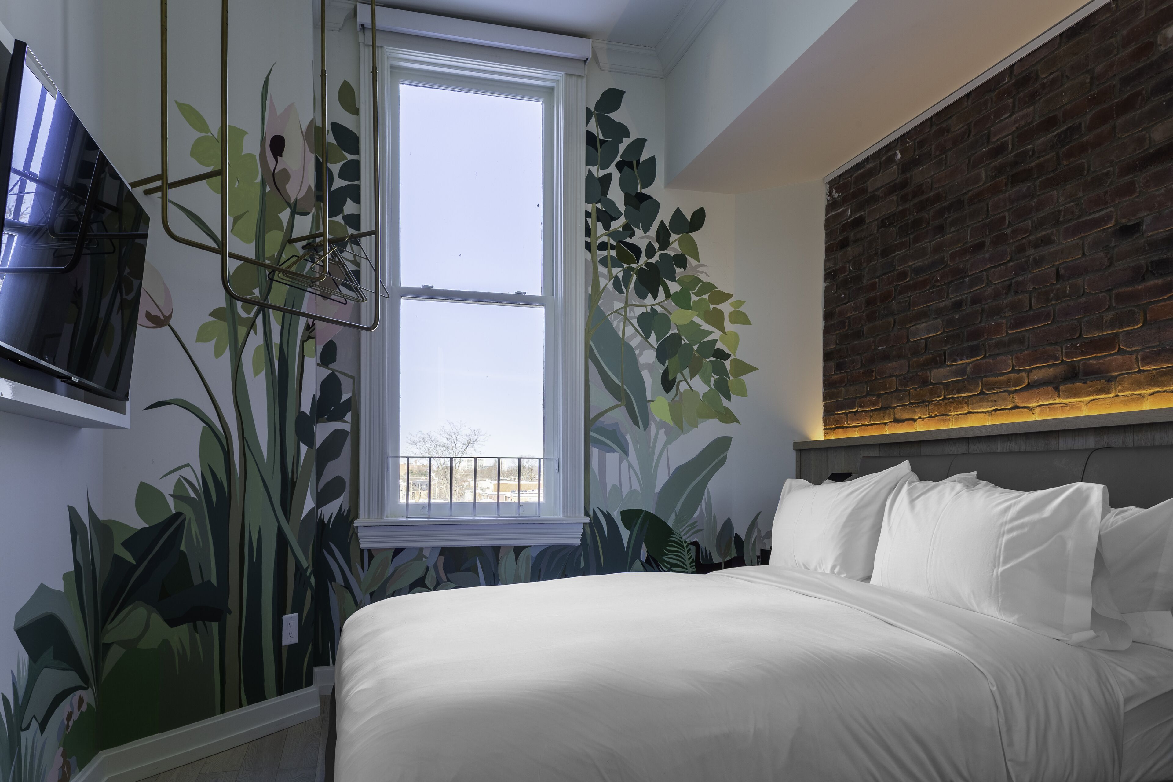 A bedroom with a large bed, a TV mounted on the wall, a window, and a brick accent wall.