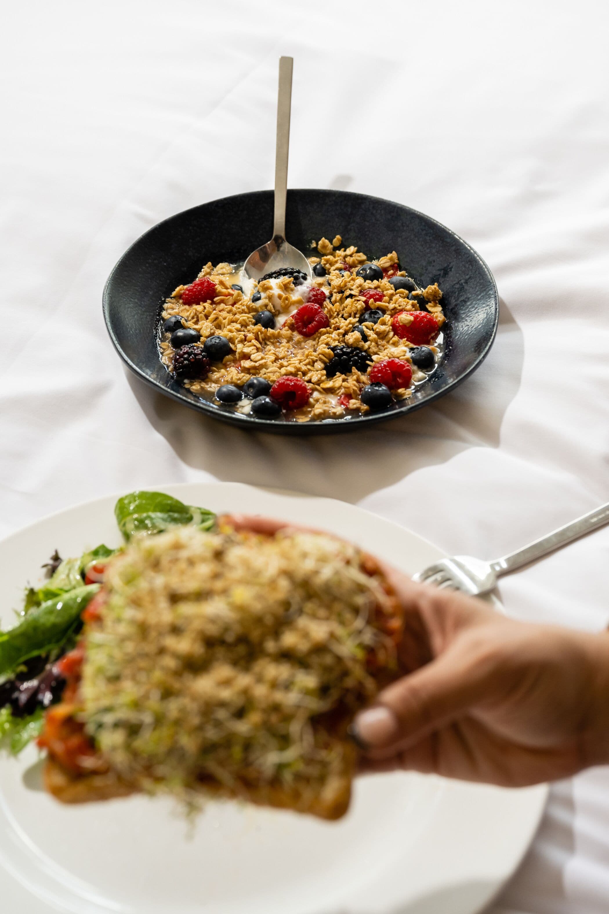 Un plato de cereal con bayas y un tenedor, y una mano sosteniendo un bocado con verduras y otro tenedor.