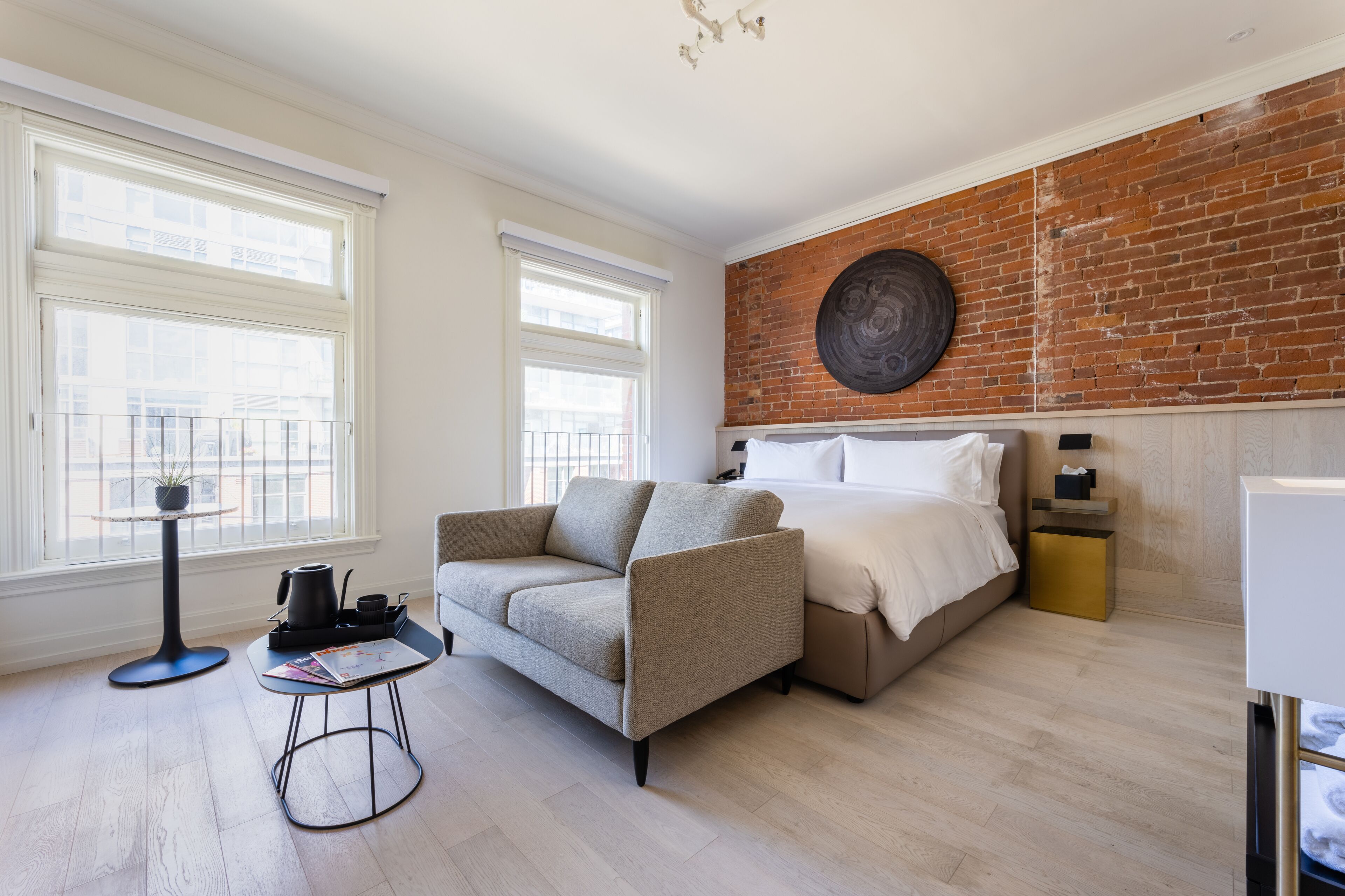 A modern bedroom with a sofa, a bed, a small table with a kettle, and a brick wall.