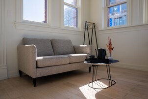 Un salon moderne avec un canapé gris, une petite table ronde dotée d’un pied noir et une fenêtre donnant sur des immeubles.