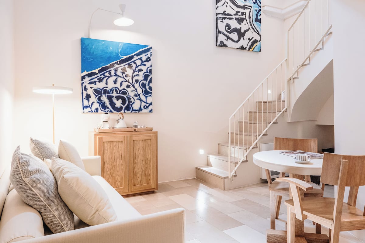 Ein modernes Wohnzimmer mit einer Couch, einem Holzschrank und einem Essbereich mit Stühlen und Tisch. Eine Treppe führt zu einer oberen Etage.