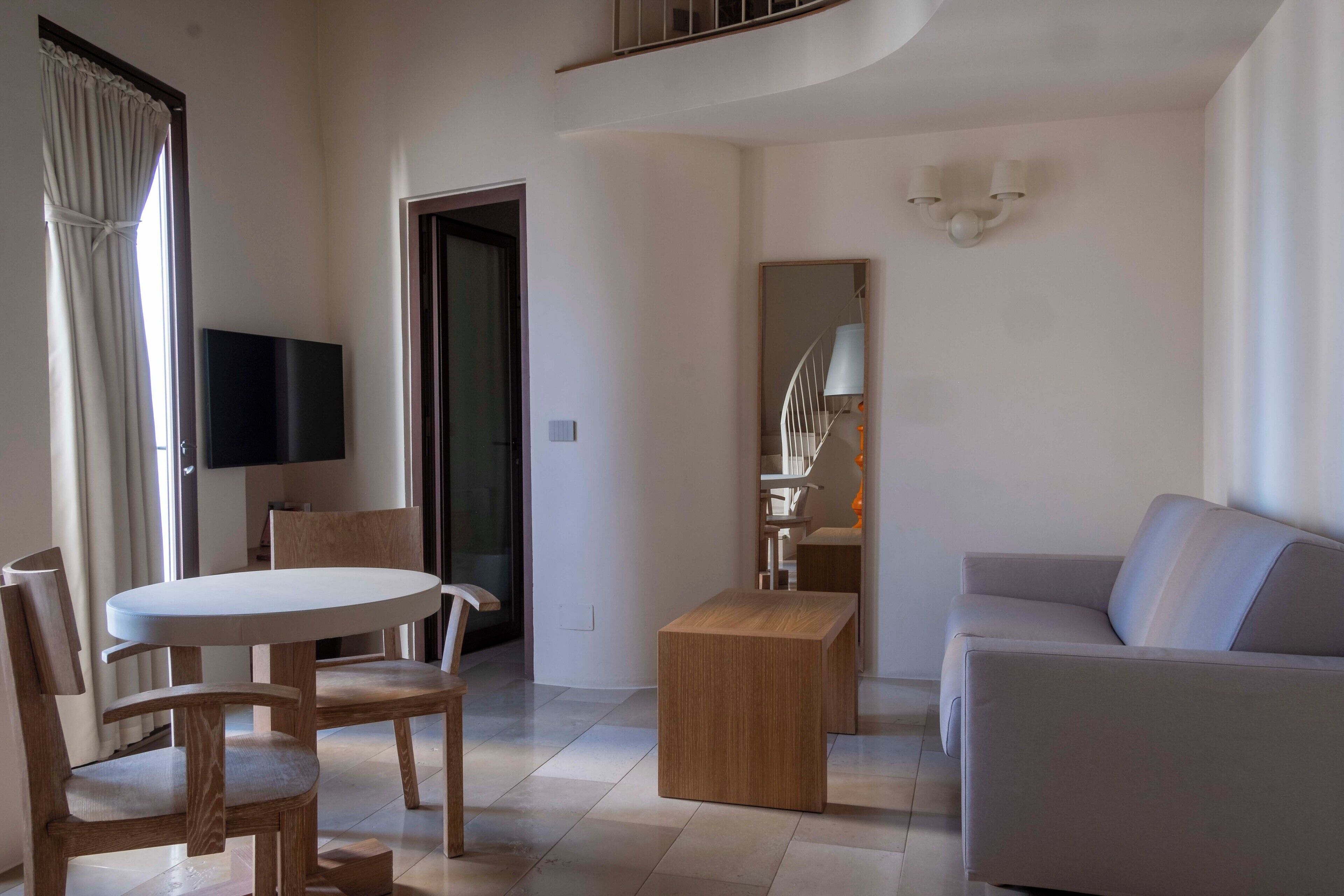 A modern living room with a round table, two wooden chairs, a sofa, a small wooden side table, and a flat-screen TV mounted on the wall.