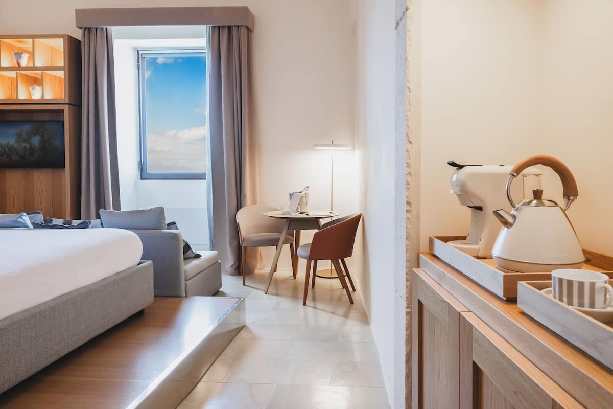 Ein modernes Hotelzimmer mit einem großen Bett, einem Schreibtisch mit Lampe, einer Kaffeemaschine und Blick zum Himmel durch ein Fenster.