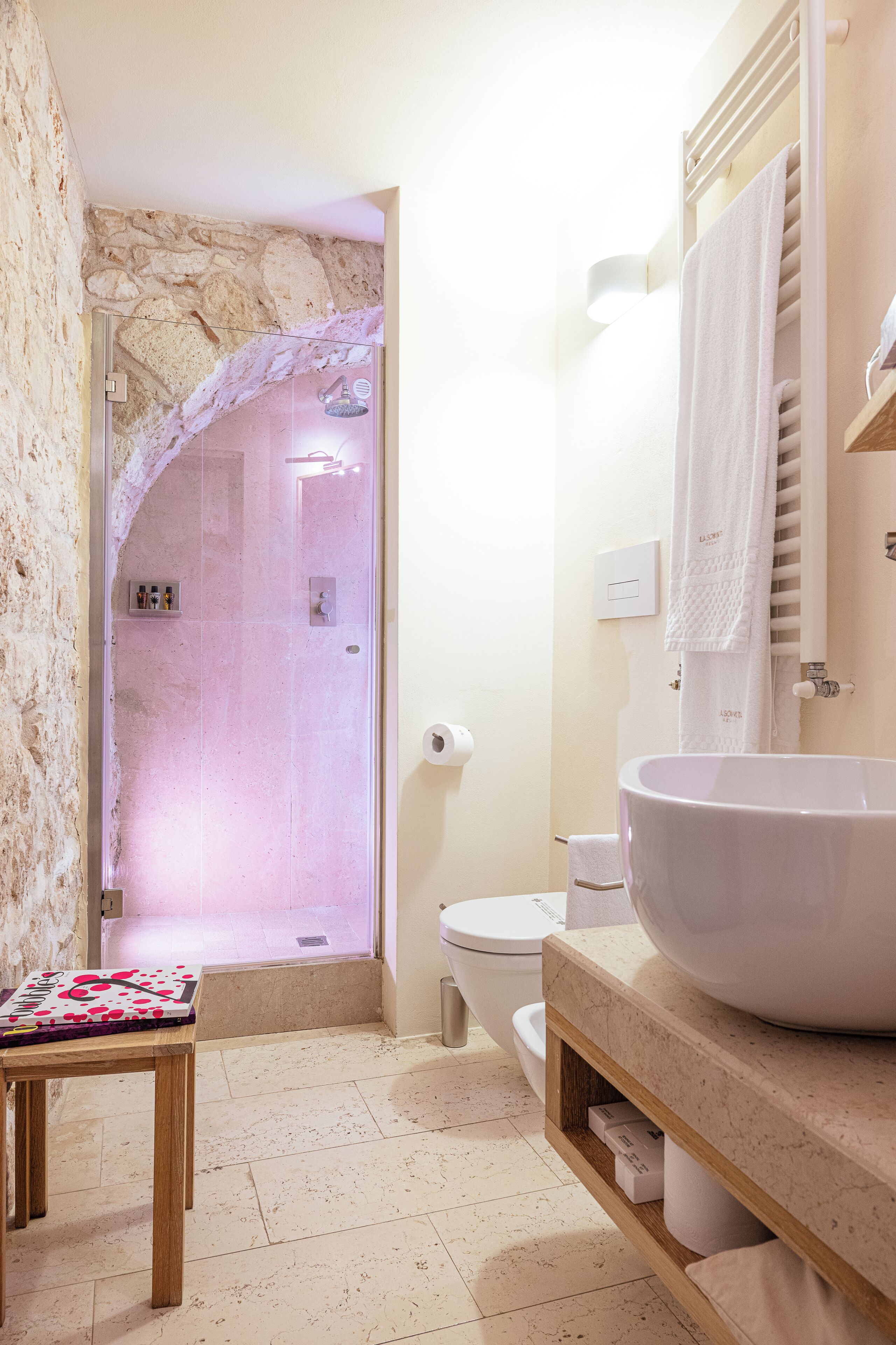 A bathroom with a stone wall, a glass-enclosed shower, a white sink, and a wooden shelf.